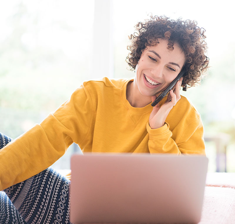 Frau mit braunen, gelockten Haaren, lachend mit einem Handy am Ohr, in gelbem Pullover vor einem Laptop sitzend. 