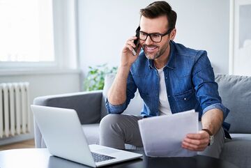 Ein Mann sitzt auf dem Sofa, telefoniert, schaut auf einen Laptop und hält Papiere in der Hand.