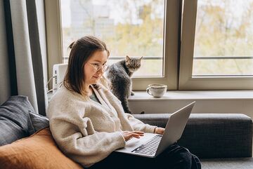 Eine dunkelhaarige Fernstudentin sitzt zuhause auf dem Sofa am Laptop, hinter ihr ihre Katze