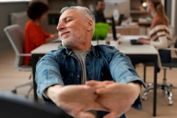 Ein Mann der zufrieden die Arme nach vorne streckt, im Hintergrund Schreibtische besetzt mit zwei Personen. 