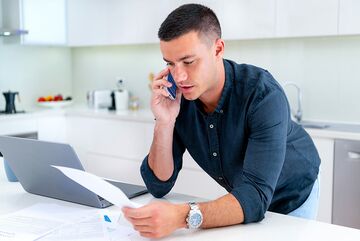 Junger Mann lehnt an Küchentisch mit Laptop, telefoniert und hält ein Paper in der Hand