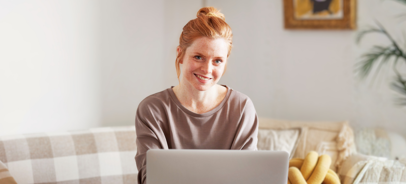 rothaarige Frau sitzt auf einem hellen Sofa mit einem Laptop und lernt