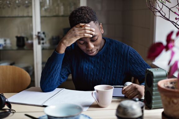 Eine junger Mann sitzt an einem Tisch und arbeitet konzentriert an einem Laptop. Neben ihr liegen geöffnete Bücher und ein Notizbuch. 