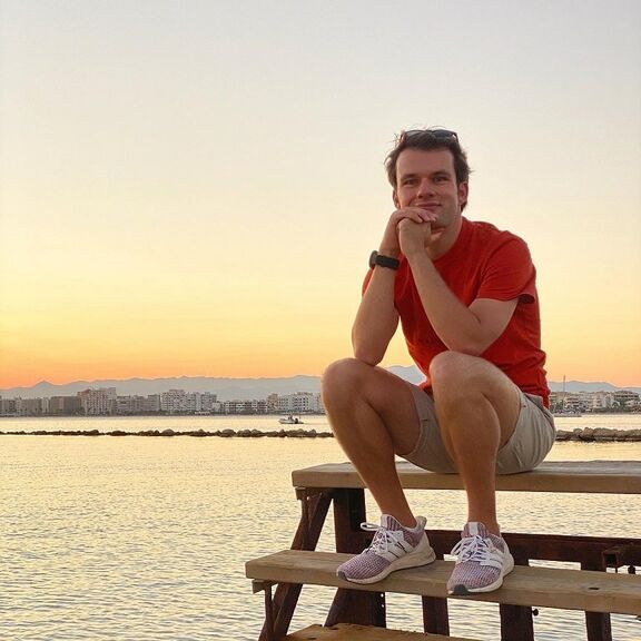 Philipp Parche sitzend in kurzen Hosen am Meer bei Sonnenuntergang