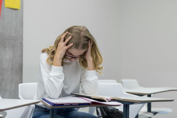 Junge Frau mit blonden langen Haaren sitzt, die Hände am Kopf, den nach unten gerichtet über Unterlagen.