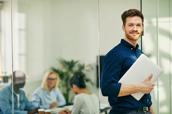 Man mit Hemd steht im Büro und hält Laptop in der Hand