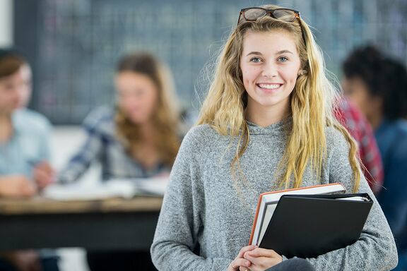 Blonde Frau mit Studienheften schaut lachend in Kamera