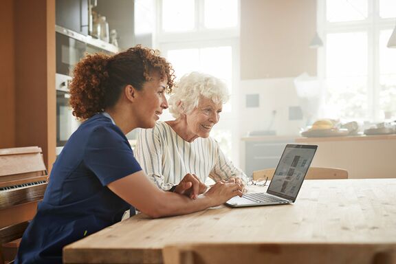 Ältere Dame und Krankenschwester sitzen gemeinsam an Tisch vor Notebook