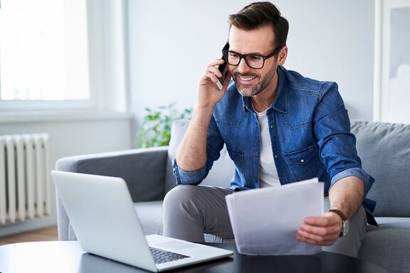 Ein Mann sitzt auf dem Sofa, telefoniert, schaut auf einen Laptop und hält Papiere in der Hand.