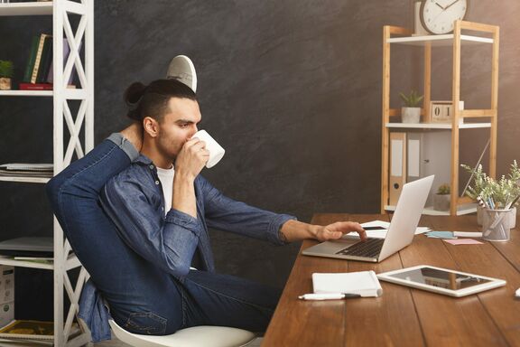 Mann sitzt vor einem Laptop, trinkt Kaffee und macht Yoga