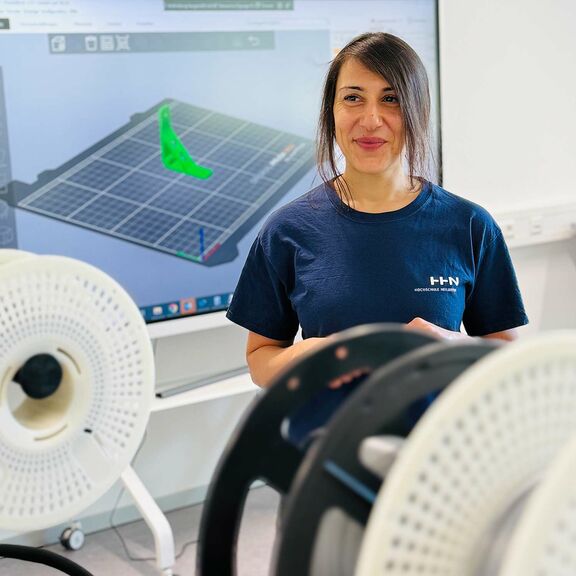 Züleyha Saraç, wissenschaftliche Mitarbeiterin, steht lächelnd vor einem Screen mit technischen Zeichnungen, im Vordergrund sind technische Geräte einer Werkstatt zu sehen.