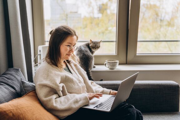 Eine dunkelhaarige Fernstudentin sitzt zuhause auf dem Sofa am Laptop, hinter ihr ihre Katze