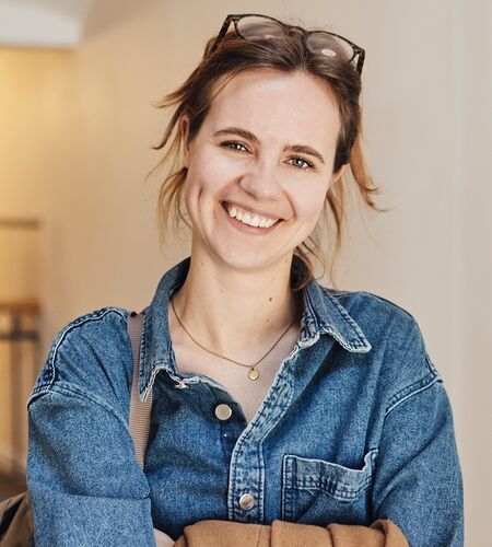 Eine junge Frau lächelt in die Kamera, im Hintergrund unscharf andere Personen im Gespräch.