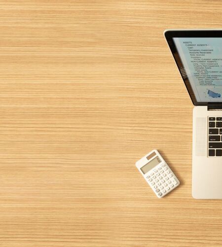 Aufgeklappter Laptop mit Statistiken auf dem Bildschirm, daneben Notizblock und Taschenrechner auf einer Holzoberfläche. 