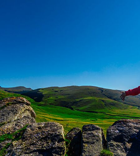 Eine Person sitzt mit dem Rücken zur Kamera auf dem Gipfel eines Berges und schaut ins Tal. Sie breitet die Arme aus und genießt die Sonne.