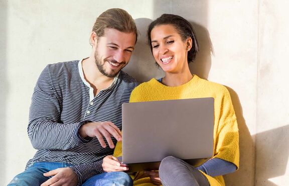 Frau und Mann sitzen an Wand gelehnt und schauen auf Notebook