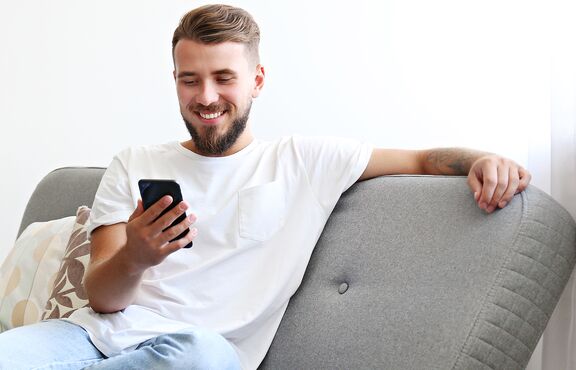 Eine lächelnde Person sitzt entspannt auf einem Sofa und hält ein Smartphone in der Hand. Die Szene vermittelt den Eindruck einer angenehmen Kontaktaufnahme von zu Hause aus, passend zum Thema Fernstudium an der HFH.