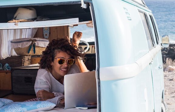 Frau liegt in Bully und schaut auf Laptop. Im Hintergrund das Meer.