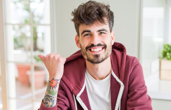 Lachender junger Mann mit Nasenpiercing und tätowiertem Unterarm in roter Joggingjacke. 