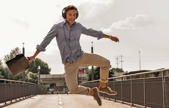 Mann auf Brücke mit Kopfhörern und Aktentasche springt freudig in die Luft