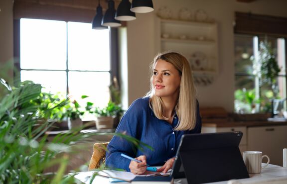Eine blonde Frau sitzt vor ihrem Laptop, macht sich Notizen und schaut verträumt zur Seite.