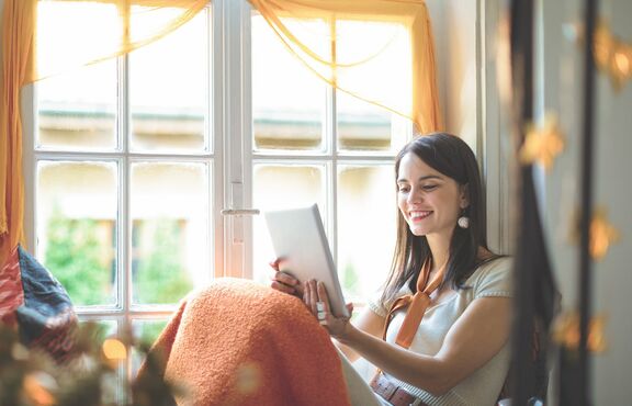 Junge Frau sitzt mit dem Rücken an einem Fensterrahmen und schaut auf ein Tablet. 