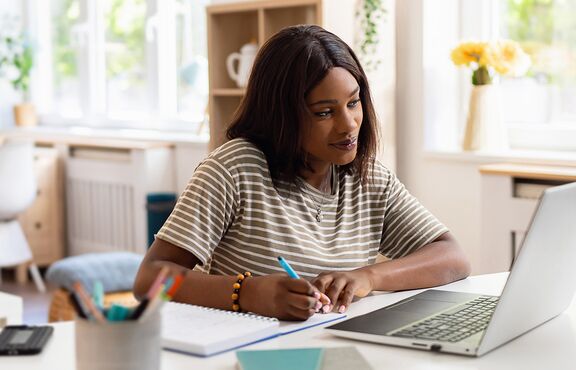 Frau sitzt lächelnd Zuhause am Laptop und macht Notizen 