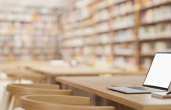 Schreibtisch mit Laptop und Büchern, im Hintergrund verschwommen dargestellte Bücherregale.