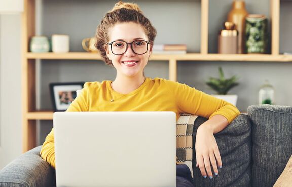 Junge Frau sitzt auf einem Sofa mit dem Laptop auf dem Schoß und blickt freundlich in die Kamera.