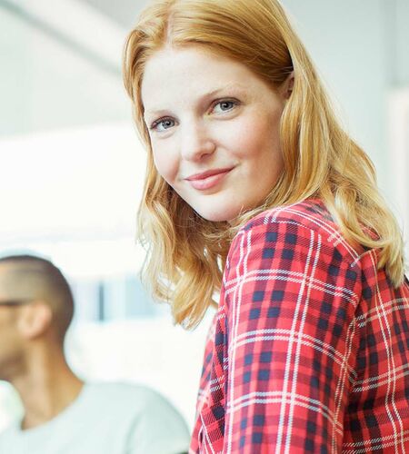 Junge Frau mit kariertem Hemd schaut freundlich in die Kamera, hinter ihr drei lernende Studierende