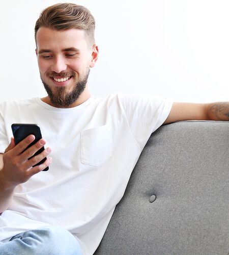 Eine lächelnde Person sitzt entspannt auf einem Sofa und hält ein Smartphone in der Hand. Die Szene vermittelt den Eindruck einer angenehmen Kontaktaufnahme von zu Hause aus, passend zum Thema Fernstudium an der HFH.