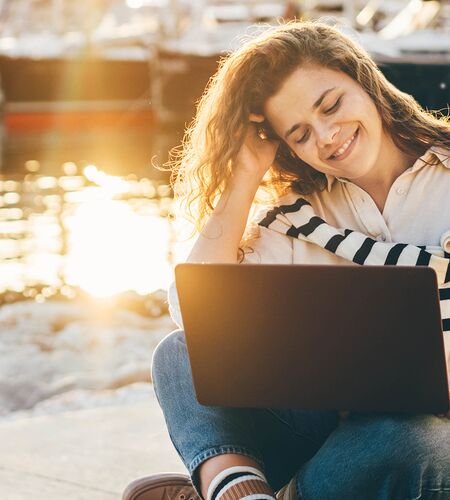 Glückliche Frau sitzt an einem Hafen und arbeitet am Laptop