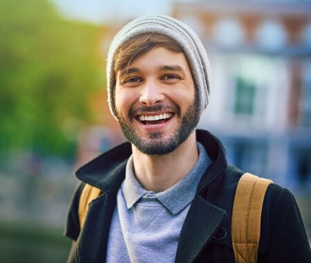 Junge Person mit weißer Mütze, schwarzer Jacke und braunem Rucksack vor unscharfem Stadt- und Grünhintergrund.