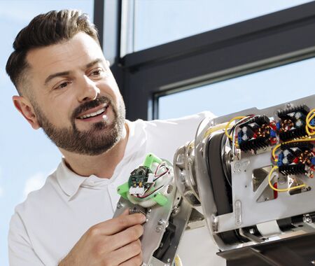 Mann in weißem Hemd arbeitet an einem komplexen mechatronischen Gerät mit sichtbaren Kabeln und Platinen vor Fenster mit blauem Himmel.