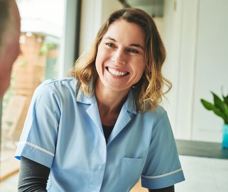 Pflegekraft in hellblauer Arbeitskleidung sitzt und spricht mit älterem Mann in brauner Jacke in hellem Raum mit Fenster und Pflanze.