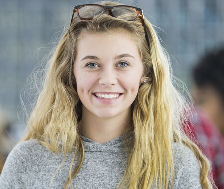 Junge Frau mit langen blonden Haaren, grauem Pullover und Brille auf dem Kopf, unscharfe Personen im Hintergrund.