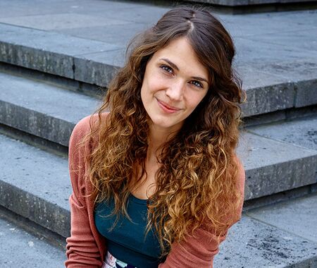 Junge Frau mit langen, lockigen Haaren sitzt auf einer Treppe aus grauem Stein und trägt ein blaues Oberteil und eine rote Jacke.