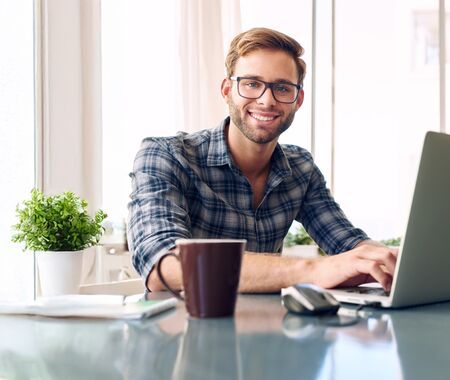 Mann in kariertem Hemd sitzt an einem Tisch mit Laptop, Kaffeetasse und Brille vor großen Fenstern mit Tageslicht.