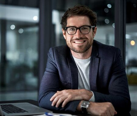 Mann in dunklem Sakko und weißem T-Shirt sitzt mit verschränkten Armen an einem Tisch mit offenem Laptop in einem modernen Büro.