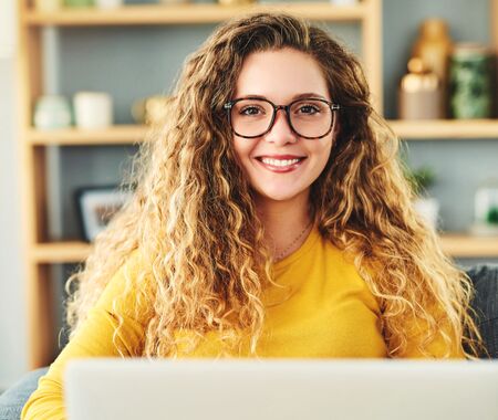 Frau mit langen, lockigen Haaren und gelbem Pullover sitzt auf einem Sofa vor einem Laptop in einem Wohnzimmer mit Bücherregal im Hintergrund.