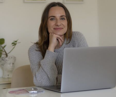 Person mit grauem Pullover sitzt an einem Tisch vor einem Laptop, im Hintergrund eine Pflanze und eine weiße Kommode.