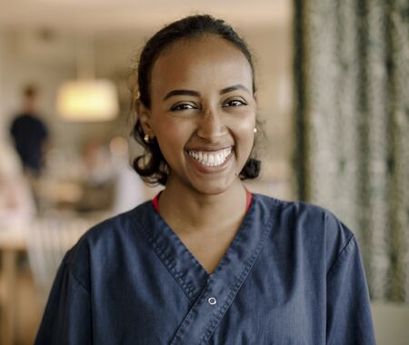 Frau in blauer Arbeitskleidung steht im Vordergrund eines Raums mit mehreren sitzenden Personen im Hintergrund.