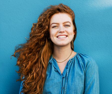 Frau mit langen, lockigen roten Haaren trägt eine blaue Bluse und steht vor einer blauen Wand.