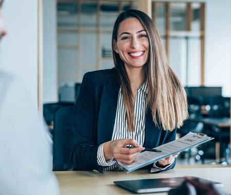 Eine Frau in Businesskleidung sitzt an einem Tisch und hält einen Stift sowie ein Klemmbrett mit Dokumenten, während sie einem Mann gegenübersitzt.