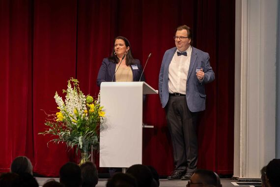 Weibliche Moderatorin und männlicher Moderator stehen auf Bühne vor Rednerpult mit Blumenstrauß daneben.