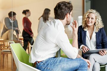 Frau und Mann unterhalten sich sitzend, im Hintergrund gehen Menschen