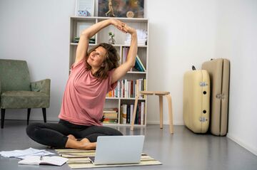 Eine Frau sitzt auf dem Boden und macht Yoga vor ihrem Laptop.