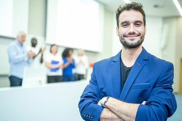Lächelnder Mann in einem blauen Sakko im Vordergrund, im Hintergrund steht eine Gruppe von Personen in einem Seminarraum