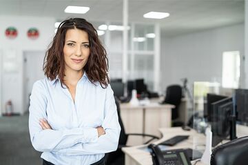 Frau mit verschränkten Armen in hellblauer Bluse steht in einem modernen Büro mit Schreibtischen und Computern im Hintergrund.