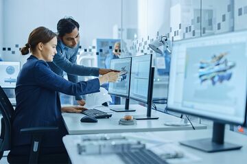 Mehrere Personen arbeiten an Computern in einem modernen Büro mit großen Monitoren und hellen Trennwänden.